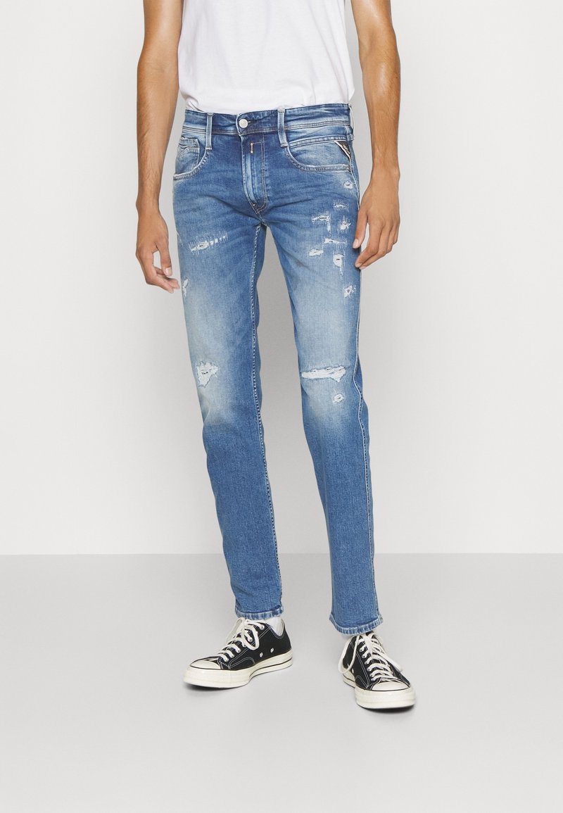 Light blue distressed denim jeans with a slim fit, featuring frayed details and a classic five-pocket design, paired with black sneakers.