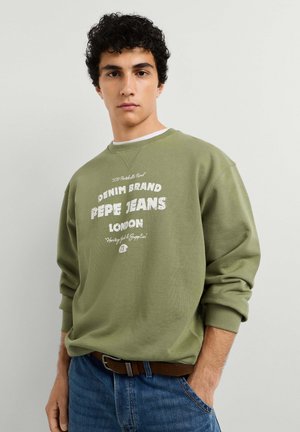 Joven con cabello rizado y oscuro que lleva una sudadera verde oliva con el texto "Pepe Jeans London" y vaqueros azules con cinturón marrón.