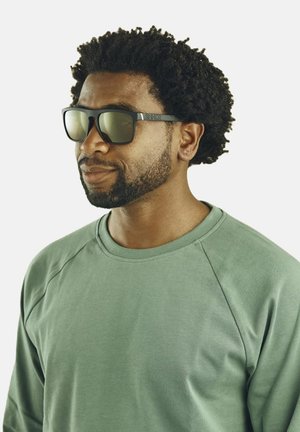 Lunettes de soleil noires avec des verres verts, dotées d'une monture noire robuste. Portées par une personne portant un sweat-shirt à manches longues de couleur vert clair.