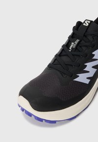 Baskets noires de sport avec une tige en maille texturée, semelle extérieure violette et un motif ondulé gris clair. Comprend des lacets et une languette à l'arrière.