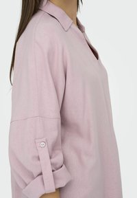 Blouse en lin rose clair avec col, décolleté en V et manches retroussées. Présente une fermeture à bouton sur les poignets pour un ajustement réglable.