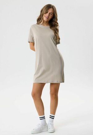 Kvinde, der står i en beige kortærmet T-shirt-kjole med hvide sneakers og hvide sokker med to sorte striber på en ensfarvet lys baggrund.