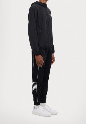 Person stående i profil iført sort hættetrøje med lynlås, sorte træningsbukser med hvide striber, hvide sokker og hvide sneakers.