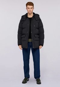 Svart vadderad jacka med huva, med en bröstficka med dragkedja, stora framfickor och en slät yta. Bärs över en svart topp och blå jeans.