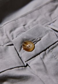 Grijze stof met een gestructureerd patroon. Een grote, ronde, bruine knoop is aan de stof vastgenaaid vlakbij een naad, met zichtbare stikdetails.