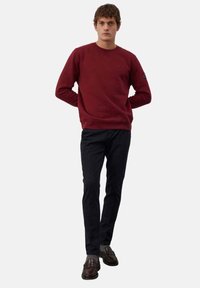 Felpa crewneck color bordeaux con polsini a coste, abbinata a pantaloni neri slim-fit e mocassini scuri. La felpa presenta un logo su una manica.