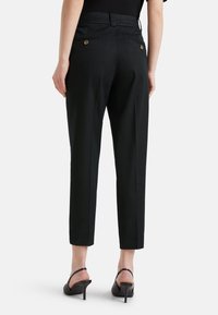 Person som bär svarta avsmalnande anklarbyxor med bakfickor med knapp och svarta klackade slingback-skor, stående mot vit bakgrund.