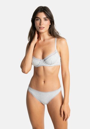 Femme aux longs cheveux bruns portant un soutien-gorge et des sous-vêtements assortis gris clair avec bordure en dentelle, debout devant un fond blanc uni.