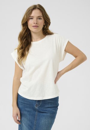 Femme aux cheveux ondulés portant des boucles d'oreilles créoles dorées, un t-shirt blanc aux manches retroussées et une jupe en denim bleu, posant avec une main sur la hanche.