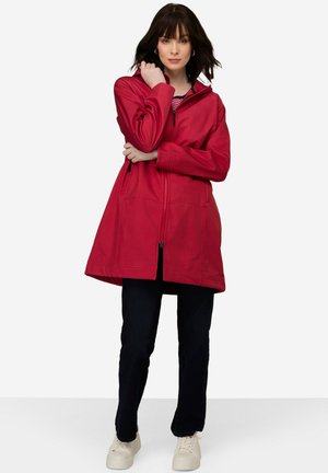 Red hooded jacket with a smooth texture, front zipper, and large pockets. Worn over dark jeans and light-colored shoes.