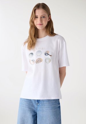 Giovane donna che indossa una maglietta bianca con design di conchiglie e la scritta "ocean", abbinata a jeans azzurri, in piedi contro uno sfondo uniforme.