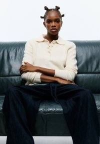 O tânăr cu coafura bantu knots stă cu brațele încrucișate pe o canapea întunecată din piele, purtând un pulover crem și pantaloni largi de culoare închisă.