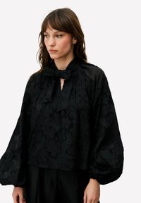 Blouse noire avec motif floral texturé, nœud papillon au col et manches volumineuses. Le matériau semble léger et légèrement transparent.
