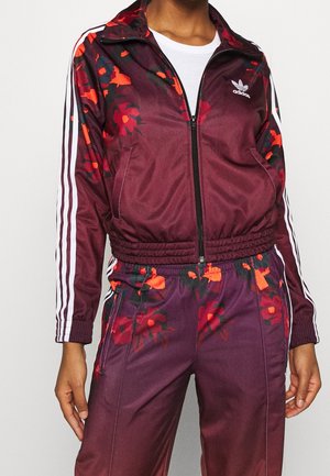 Personne portant un survêtement Adidas marron rouge avec des motifs floraux rouges, des bandes blanches sur les manches et les pantalons, et une veste blanche zippée par-dessus une chemise blanche.