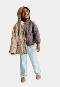 Quiltet jakke med en brun side og en beige side med et linjemønster, hætte og lynlås; parret med lyseblå jeans og brune sko.