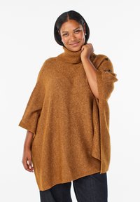 Brązowy dziany ponczo z golfem, prążkowaną fakturą i guzikami na rękawach, noszony na ciemnych dżinsach.