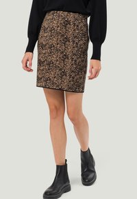 Jupe noire, ajustée, arrivant au genou avec un motif texturé de touches dorées. Portée avec des bottines noires et un pull noir.