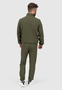Olijfgroene trainingspak met een ritsjack en bijpassende broek met zijstreepjes, gemaakt van een soepele, flexibele stof. Witte sneakers maken de look af.