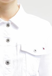 Camicia bianca in cotone con colletto, bottoni argentati e un piccolo dettaglio del logo sulla tasca. Texture liscia.
