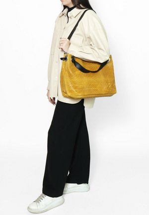 Borsa gialla intrecciata con un motivo testurizzato, dotata di una tracolla nera e di un design spazioso, tenuta da una persona in abbigliamento casual.