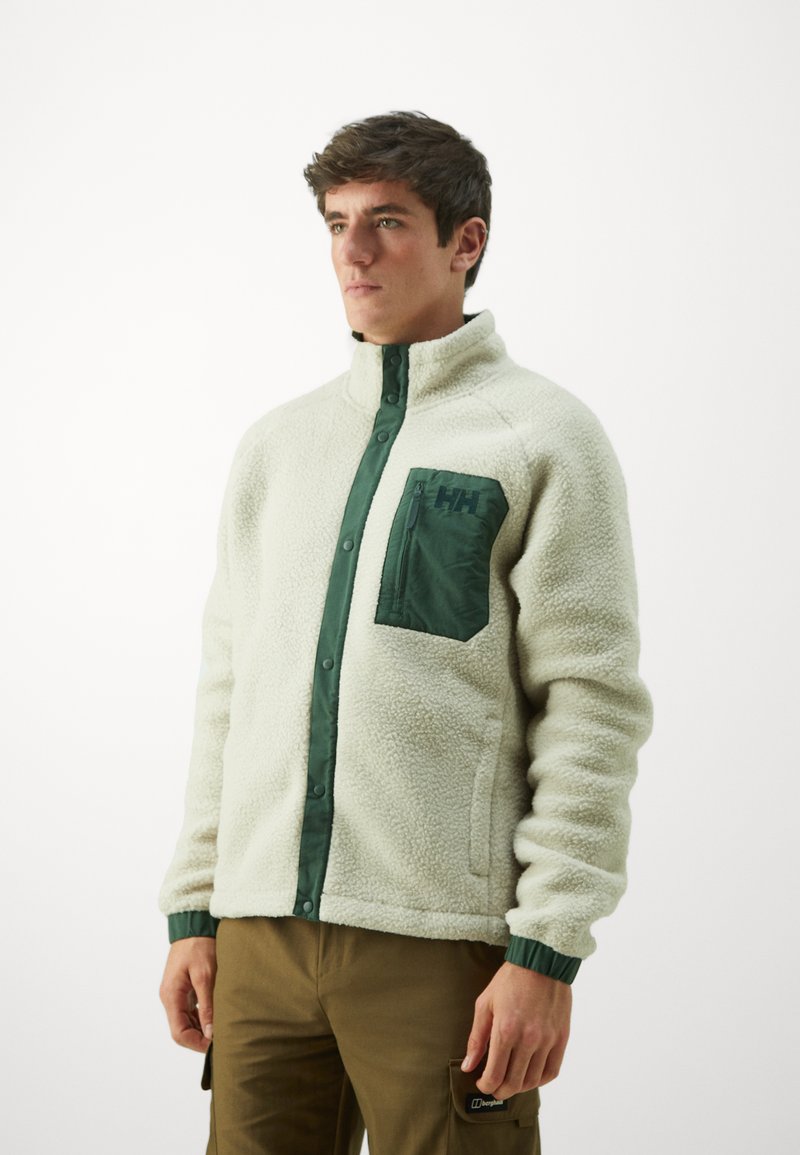 Chaqueta de forro polar en color blanco roto con un bolsillo en el pecho de color verde con cremallera, cuello alto y acentos verdes en los puños y el tapón frontal.