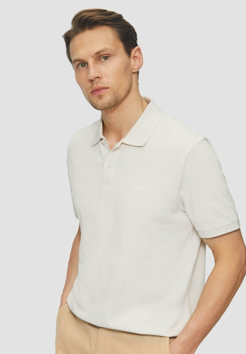 Licht crèmekleurig poloshirt van zachte stof, met een kraag en drie knopen. Subtiele logo-detail op de borst.