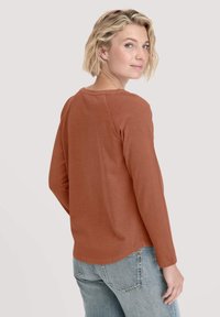 Langärmliges, rostfarbenes Oberteil aus weichem Stoff mit rundem Ausschnitt und genähten Details, kombiniert mit hellblauen Jeans.