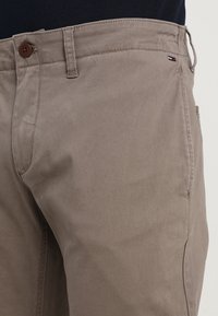 Beżowe bawełniane spodnie o gładkiej fakturze, z brązowym guzikiem, kieszeniami po bokach oraz małą naszywką z logo w kolorach czerwonym, białym i niebieskim na pasku.