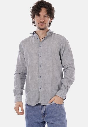 Hombre con cabello rizado que lleva una camisa de botones gris claro con rayas verticales y jeans azules, de pie con una mano en el bolsillo.