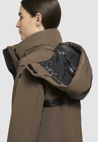 Giacca imbottita marrone con colletto alto, fodera interna nera e dettagli con logo sulla patta. Presenta un esterno liscio e texture.