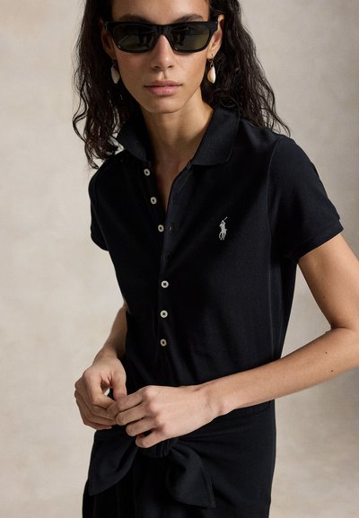 Žena v čiernej zapínanej polo košeli s logom a s mušľovými náušnicami, upravuje viazanú látku v páse, na neutrálom pozadí.