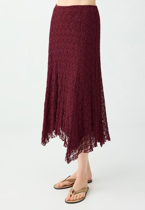 Femme portant une jupe midi en dentelle bordeaux avec un ourlet asymétrique et des sandales beige type tongs sur un fond blanc uni.