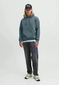 Felpa con cappuccio in blu-grigio tenue, misto cotone, con cordini regolabili. Abbinata a jeans grigio scuro e sneakers beige. Cappellino nero.