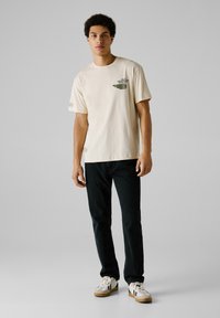 Camiseta de algodón crema con mangas cortas, gráficos impresos en verde oscuro en el pecho, combinada con jeans negros y zapatillas blancas.