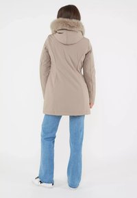 Giacca impermeabile beige con cappuccio foderato in pelliccia, silhouette aderente e texture liscia. Abbinata a jeans a zampa d'elefante blu chiaro e sneakers nere.