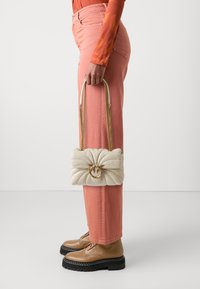 Kremo spalvos siuvinėta rankinė su auksine grandine ir apvaliu detaliu, laikoma asmens, apsirengusio rožinėmis kelnėmis ir rudais batais.