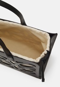 Sac fourre-tout en cuir noir avec un design floral découpé, doublure en tissu beige, texture douce et poignées en cuir. Détails de la fermeture à cordon visibles.