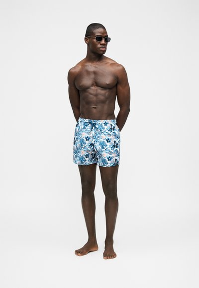 Άντρας χωρίς πουκάμισο, φορώντας μαύρα γυαλιά ηλίου και μπλε μαγιό με λουλουδάτο σχέδιο, στέκεται ξυπόλητος μπροστά σε ένα απλό φωτεινό φόντο.