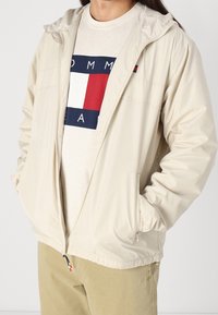 Lehká světle béžová bunda s kapucí, vybavená zipem vpředu a elastickými manžetami. Kombinována s krémovým tričkem a khaki kalhotami.