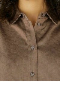 Camicia marrone a trama con colletto e quattro bottoni, indossata da una persona con capelli ricci scuri visibili sulle spalle.