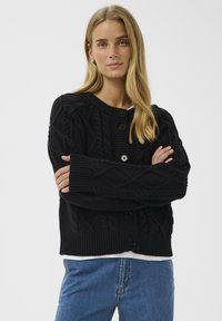 Fiatal nő, hosszú szőke hajjal, fekete kötött gombos kardigánt és kék farmert viselve, karba tett kézzel áll.