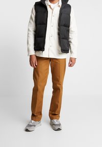 Man som bär en svart pufferväst över en vit skjorta, bruna byxor och grå träningsskor står mot en ren vit bakgrund.