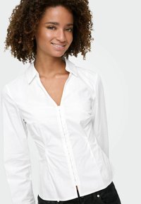Blouse en coton blanc à coupe ajustée, manches longues et col en V. Dotée d'une fermeture à boutons et d'une texture lisse.