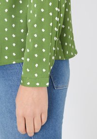 Blusa verde con estampado de puntos irregulares blancos, mangas largas y dobladillo recto, combinada con jeans de mezclilla azul.