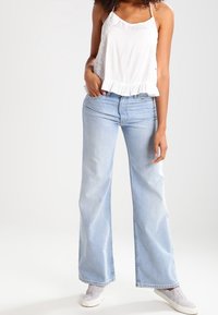 Top blanco sin mangas con dobladillo con volantes y ribete de encaje, combinado con pantalones de mezclilla de pierna ancha de color azul claro. La modelo lleva unos zapatos gris claro sin cordones.