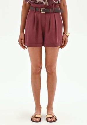 Personne portant un short bordeaux taille haute avec une ceinture foncée, un haut à motifs, un bracelet doré et des sandales plates, debout sur fond blanc.