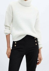 Pull en maille côtelée crémeux à col roulé associé à un pantalon noir taille haute avec des accents de boutons dorés. Texture douce et coupe ajustée.