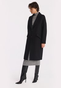 Kobieta w długim czarnym płaszczu z guzikami, nosząca szary sweter typu golf i czarne kozaki na wysokim obcasie do kolan, stoi na białym tle.