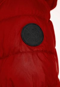 Doudoune rouge avec finition mate, texture matelassée et un patch logo en caoutchouc noir avec des détails en relief sur le côté gauche.