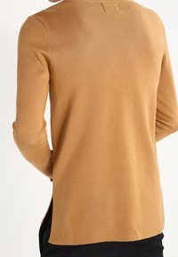 Jasnobrązowy sweter z dzianiny z długimi rękawami, okrągłym dekoltem i bocznymi rozcięciami. Tekstura wydaje się miękka i lekko prążkowana.